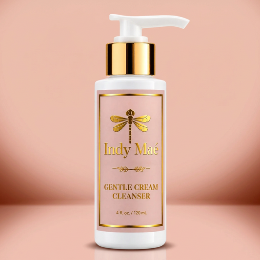 A bottle of Indy Mae Gentle Cream Cleanser with a white pump dispenser, a gold collar, and a light pink label with gold lettering, set against a solid pale pink background.