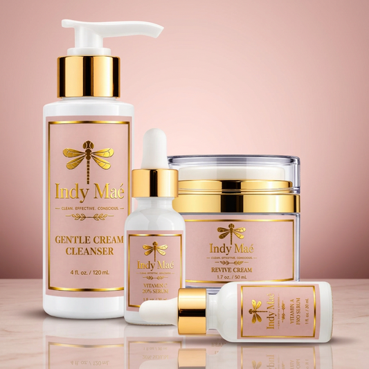 A set of five skincare products from Indy Maé on a solid pale pink background. The products include a Gentle Cream Cleanser pump bottle, a Revive Cream jar, a Vitamin C serum dropper bottle, and two smaller ampoules, all featuring white bottles with shiny gold caps and labels with a gold dragonfly logo.