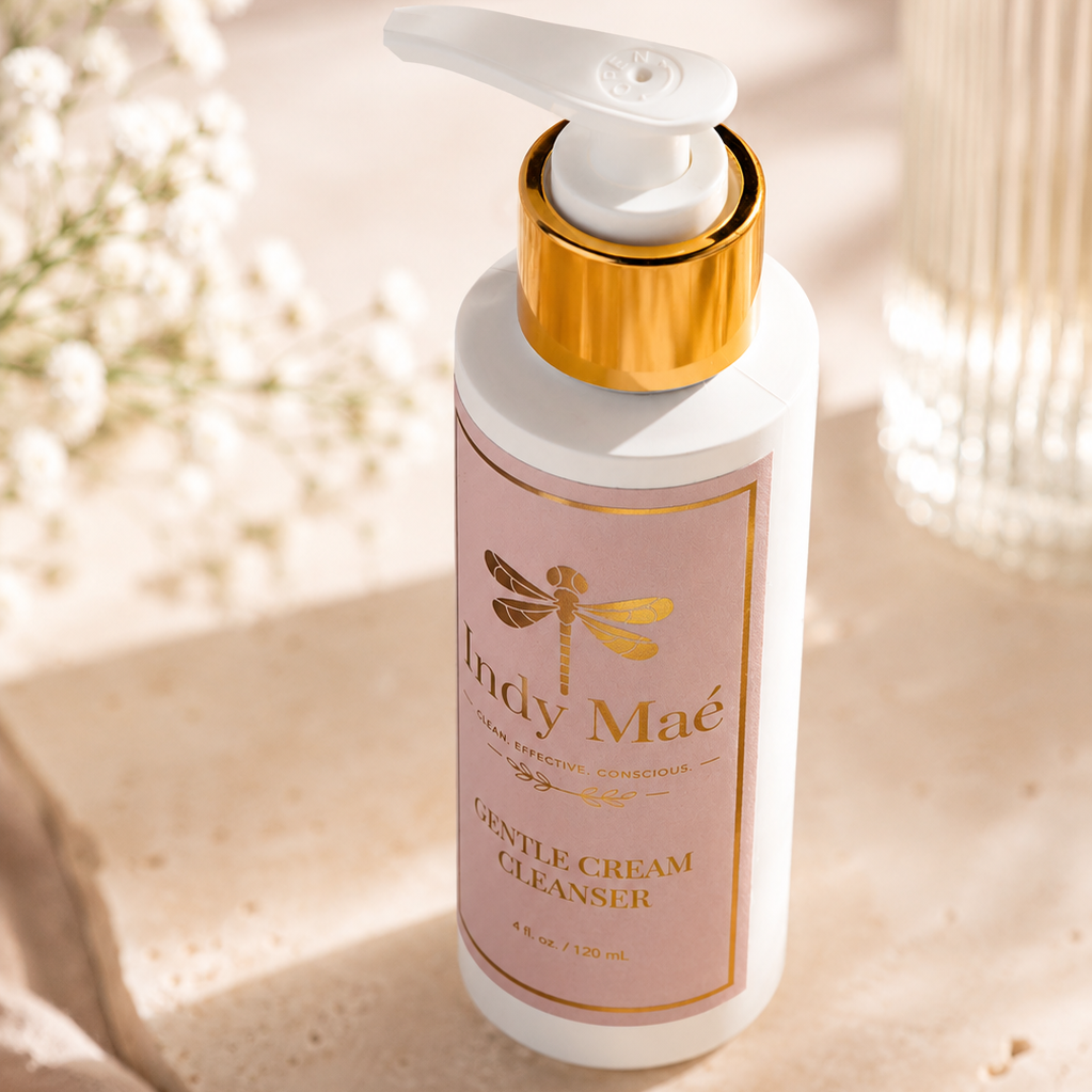 A bottle of Indy Maé Gentle Cream Cleanser with a white pump and gold collar, set on a textured beige stone surface with soft floral props in a warm, sunlit setting.