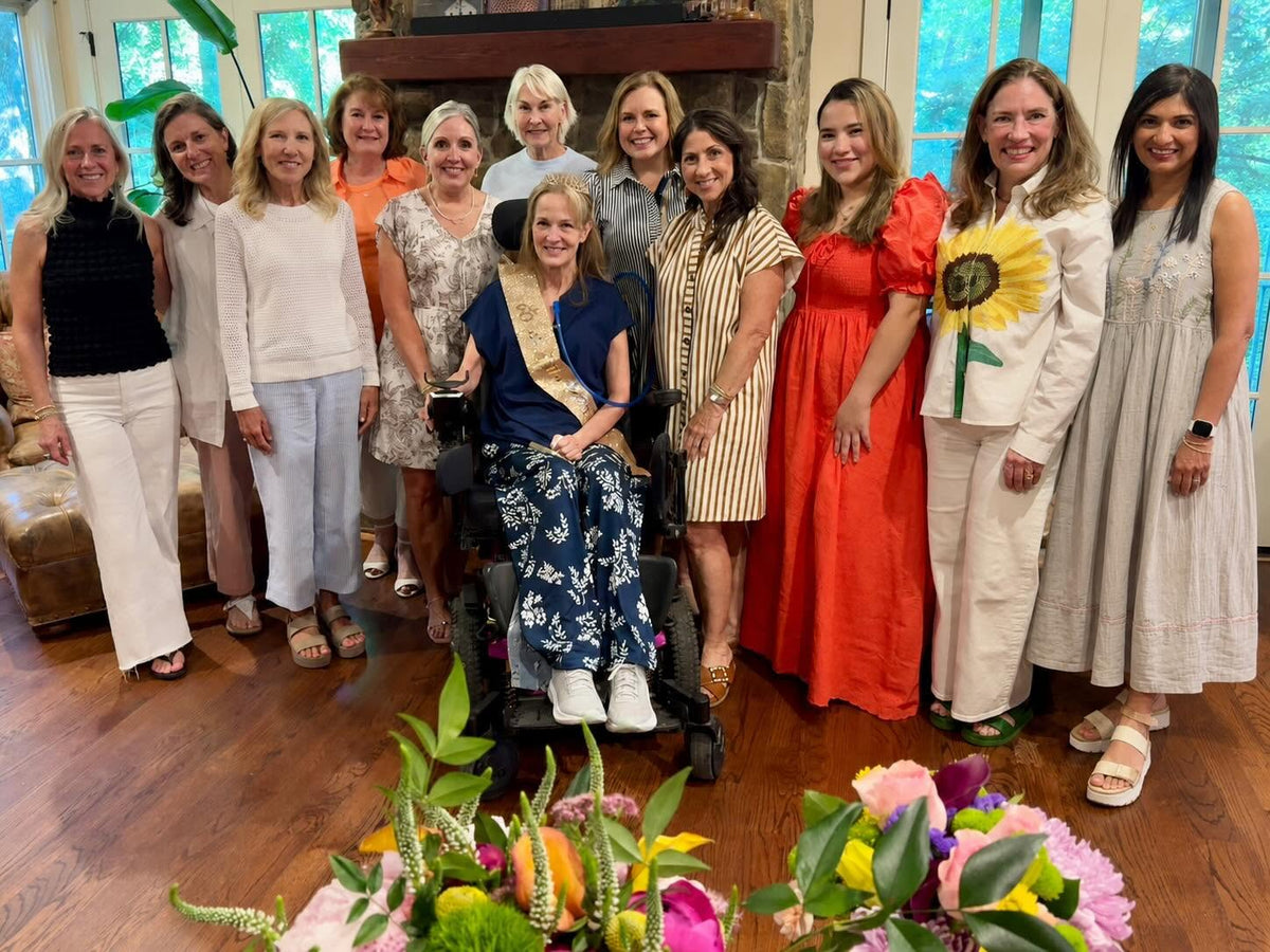Group of women posing for a photo with a woman in a wheelchair, surrounded by flowers and plants.